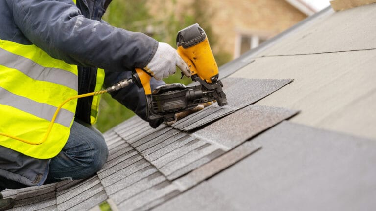 Worker installing shingles on a roof with nail gun.